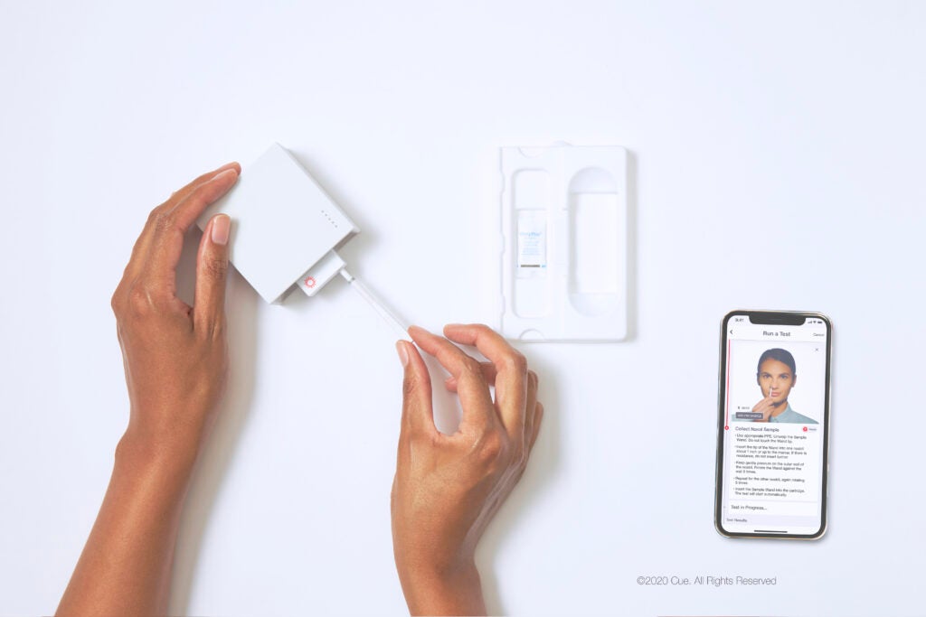 top down view of a pair of hands using a white square reader, inserting a white stick into the reader. on the right is a smartphone of a person demonstrating instructions for an at home covid test