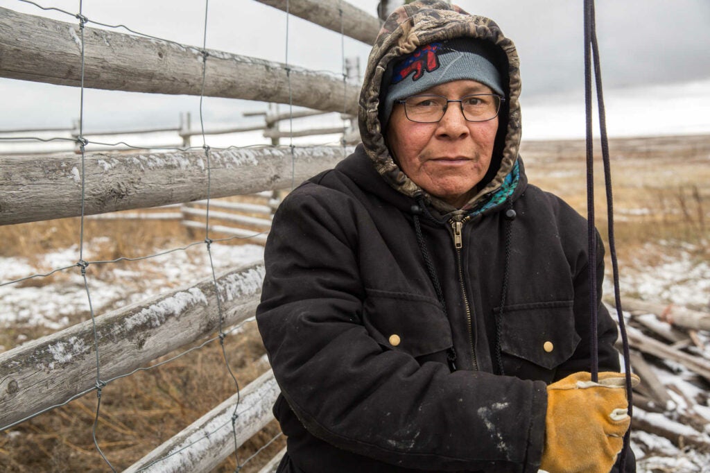 Bison rancher Man Black Plume standing in front of cattle fencing in a black jacket and work gloves