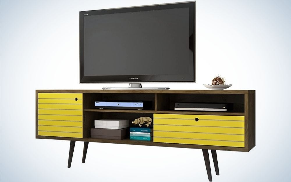 El televisor negro se encuentra en un moderno mueble de TV con patas rociadoras con tres estantes.  Dos de ellos son estantes de madera amarilla, un armario y un cajón.