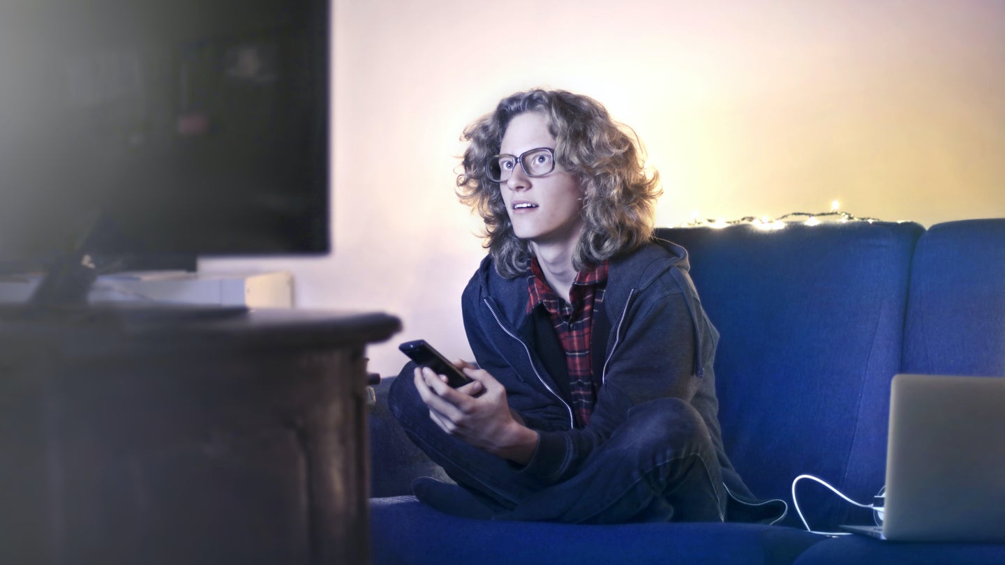 A person with curly shoulder-length blonde hair sitting on a blue couch and leaning forward to watch TV while holding a TV remote in their hands.