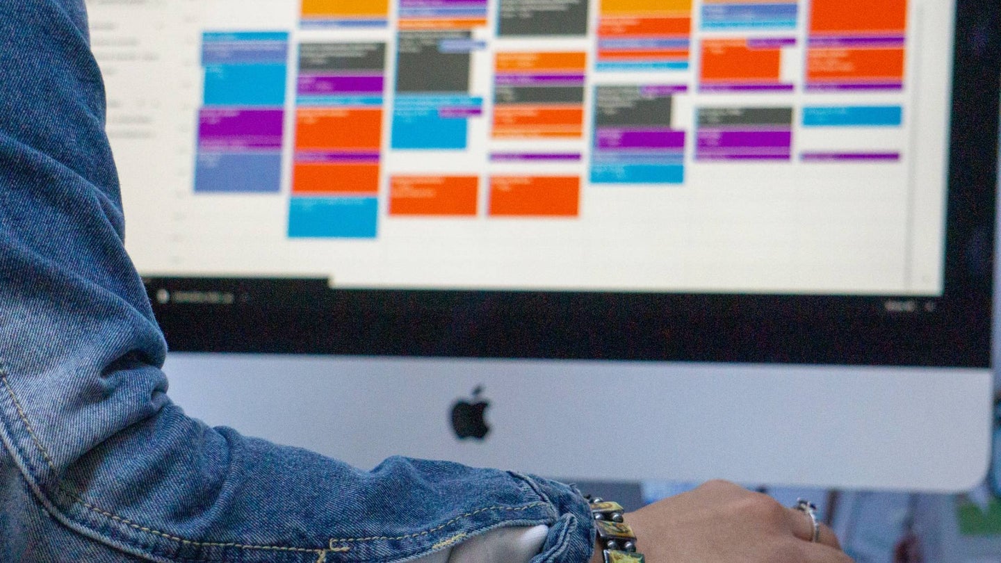 A person wearing a denim jacket using a very full Google Calendar on an Apple desktop computer.