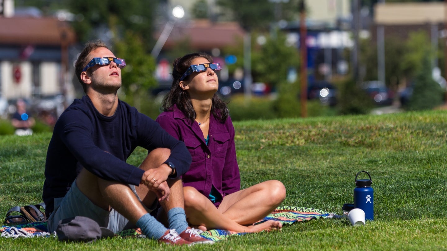 a couple wearing glasses sits on the grass watching the eclipse