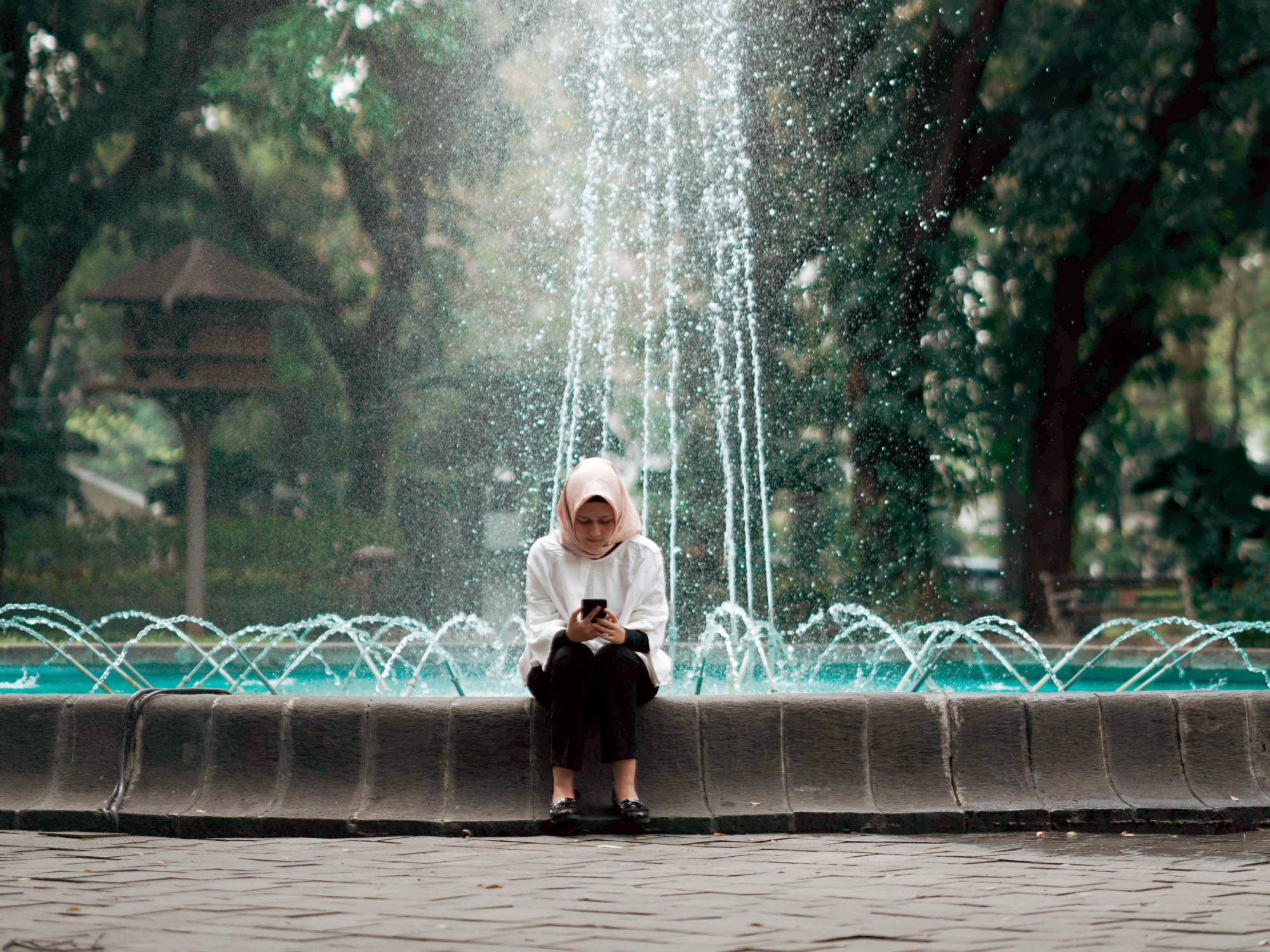 A woman sitting at the edge of an ornate park fountain, looking at a phone.