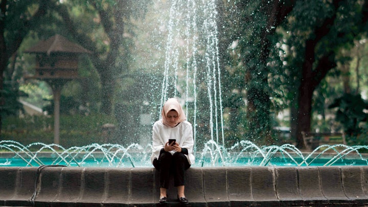A woman sitting at the edge of an ornate park fountain, looking at a phone.