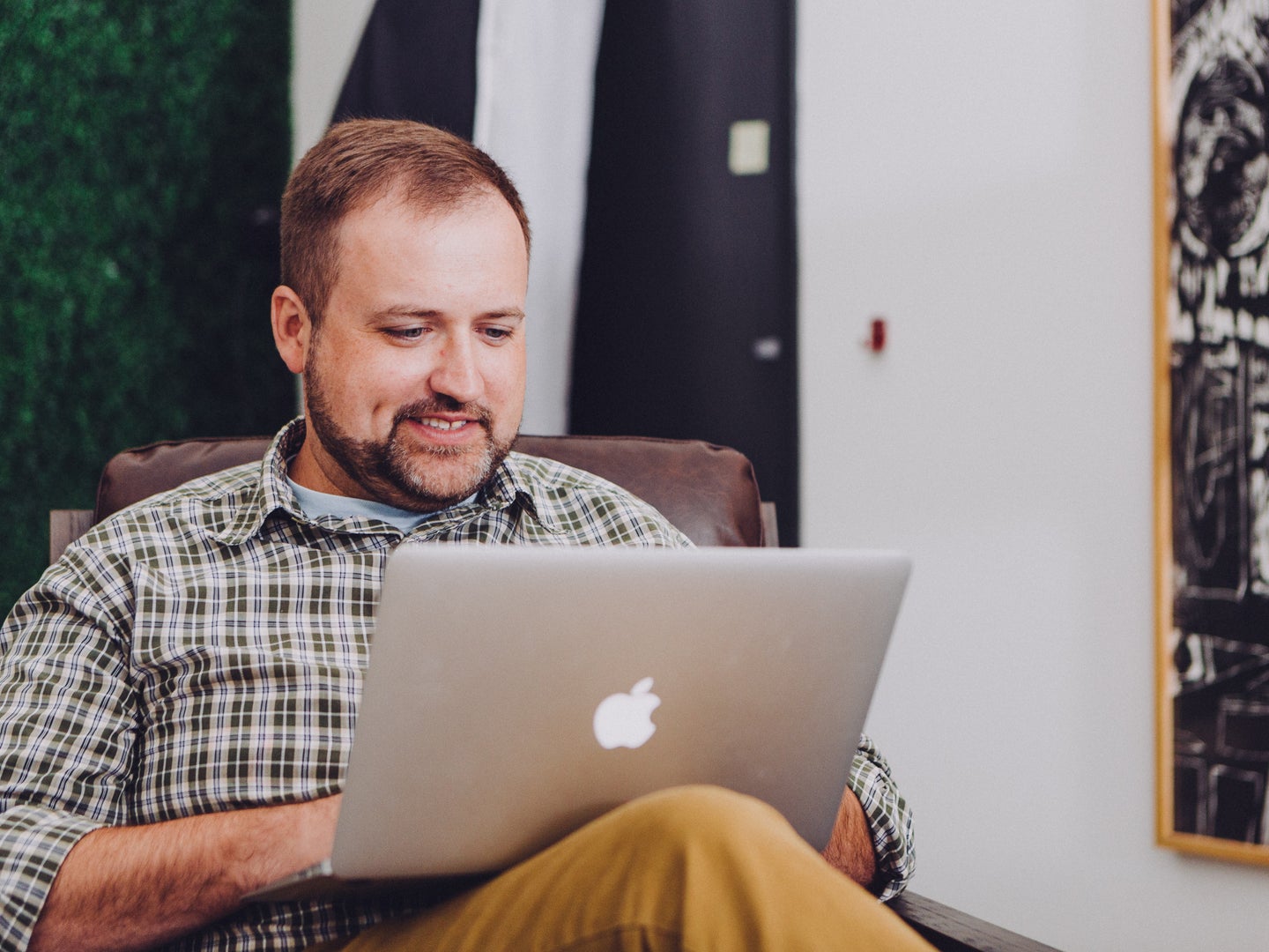 A man in a green plaid shirt sitting in a chair and smiling while using a Macbook laptop.