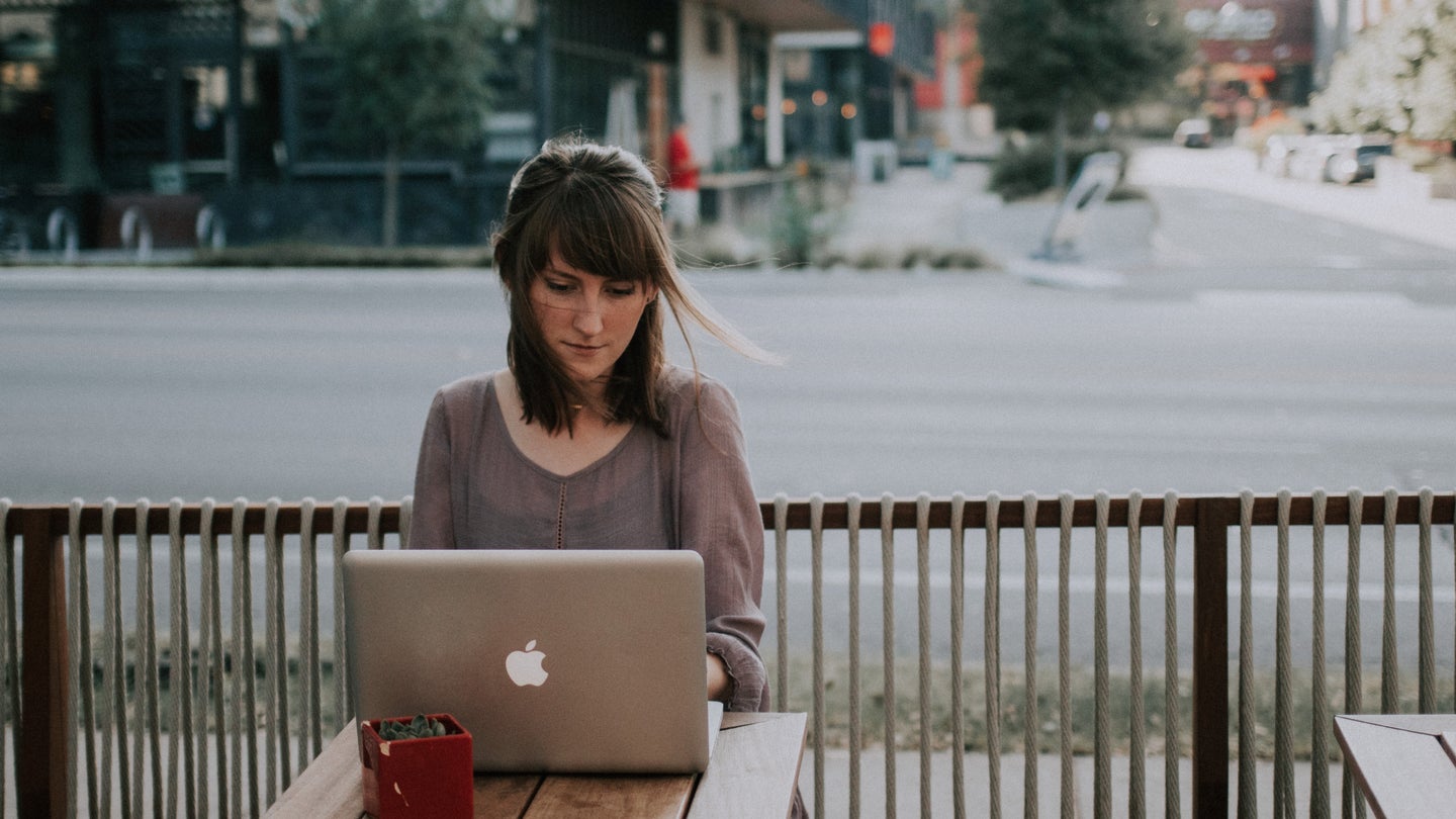 A young woman sitting outside a coffee shop on a patio by the street, using a silver Macbook on a wooden table.