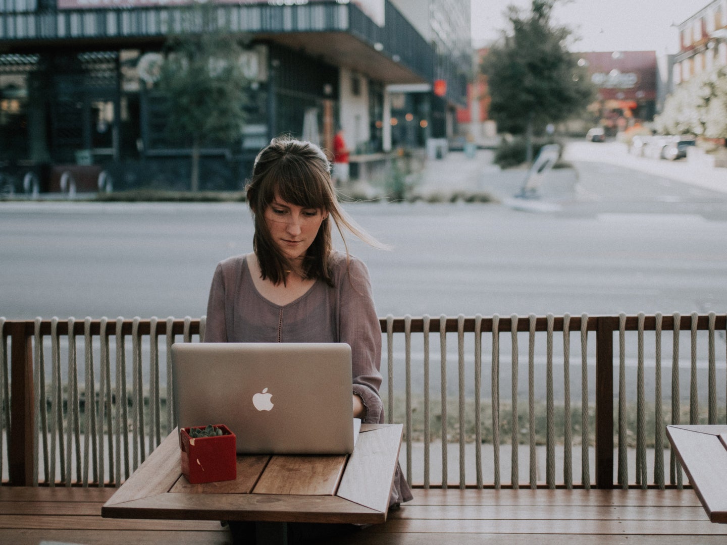A young woman sitting outside a coffee shop on a patio by the street, using a silver Macbook on a wooden table.