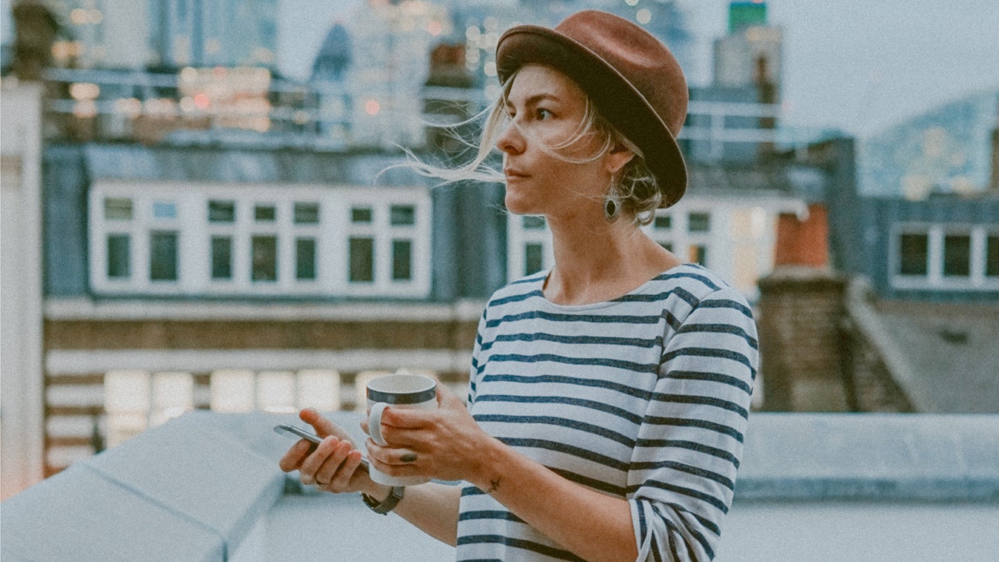 A woman wearing a brown hat and a striped shirt, standing on a rooftop during the day and holding a coffee and her phone.