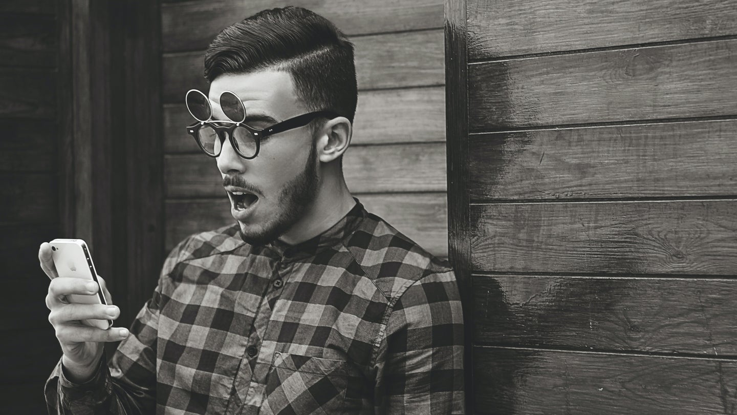 A young man in a plaid shirt standing against a wooden wall wearing glasses with flip-up outdoor lenses, looking at his phone with a shocked face.