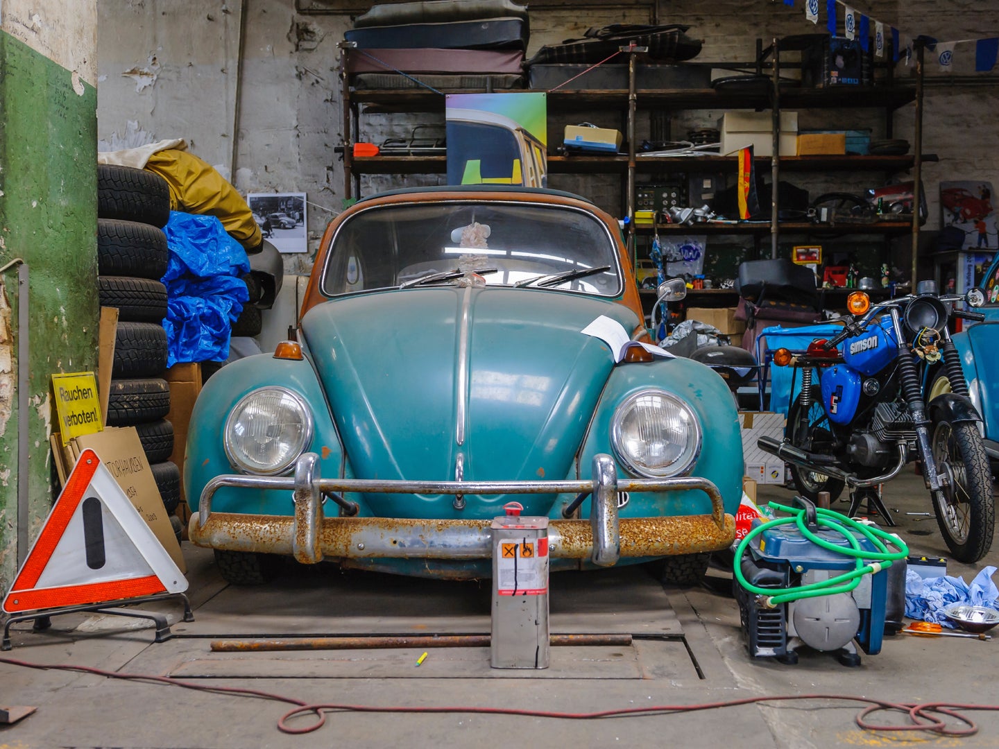 An old Volkswagen Beetle in a garage with a motorcycle and a lot of clutter.