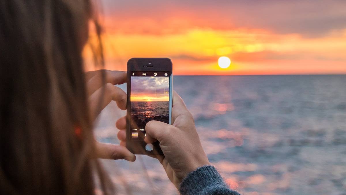 A woman standing by the ocean, using her phone to take a photo of the sunset over the water.