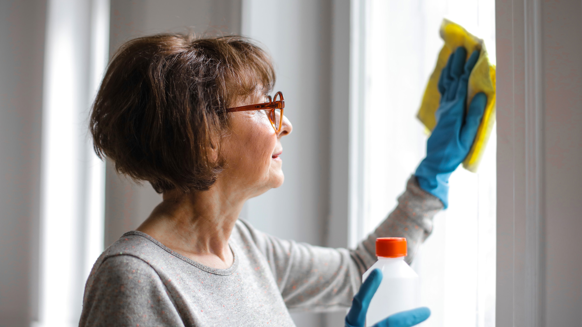 An older person with short brown hair wearing blue rubber gloves and cleaning a window with a yellow rag.