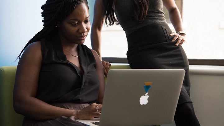 A woman using an Apple Macbook laptop while another woman watches from nearby.