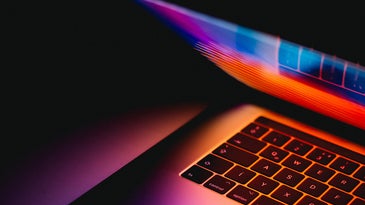 A partially closed Macbook laptop emitting multi-colored light in a dark room.