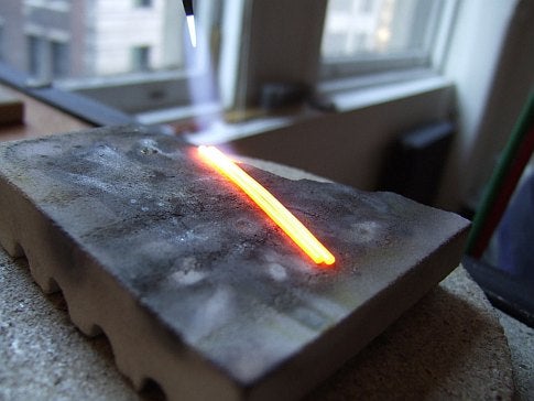 A red-hot strip of metal on an anvil near a window.