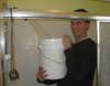 A clothed man standing in a shower and filling a white 5-gallon bucket from the showerhead.