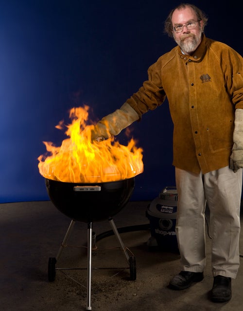 A man with short gray hair and a beard wearing protective gear and standing next to a charcoal grill fire.