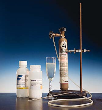 A canister of chlorine gas to the right of a champagne glass full of hydrogen peroxide. There are two white bottles on the left, one of bleach and one of peroxide.