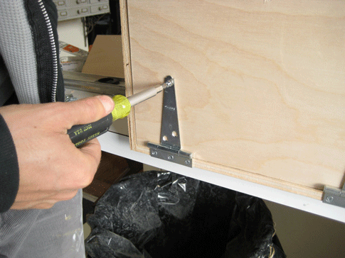A person using a screwdriver to attach hinges to a plywood newspaper baler.