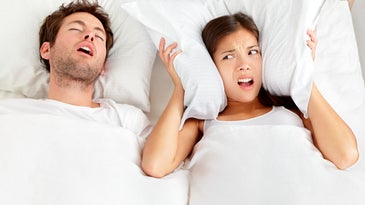 A man snoring in a white bed, while a woman angrily holds a pillow over her ears next to him.
