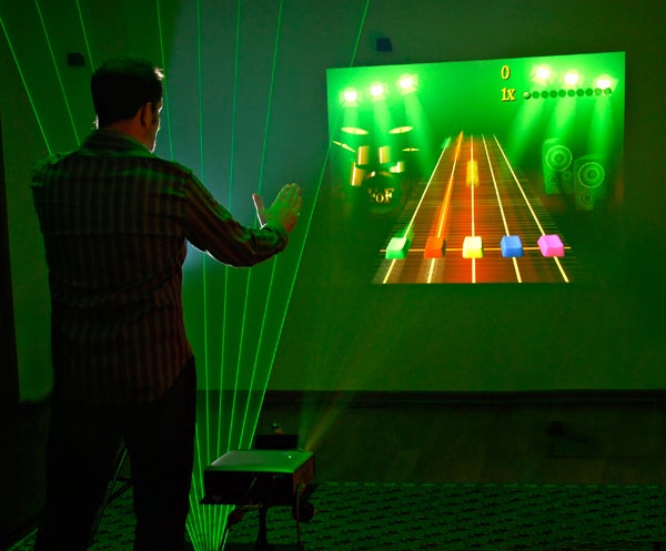 A man standing in front of a harp with strings made out of green lasers, playing Guitar Hero on a visual projected onto a green wall.