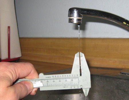 A sink faucet with a thin stream of water coming out of it and a person measuring the water's width with calipers.