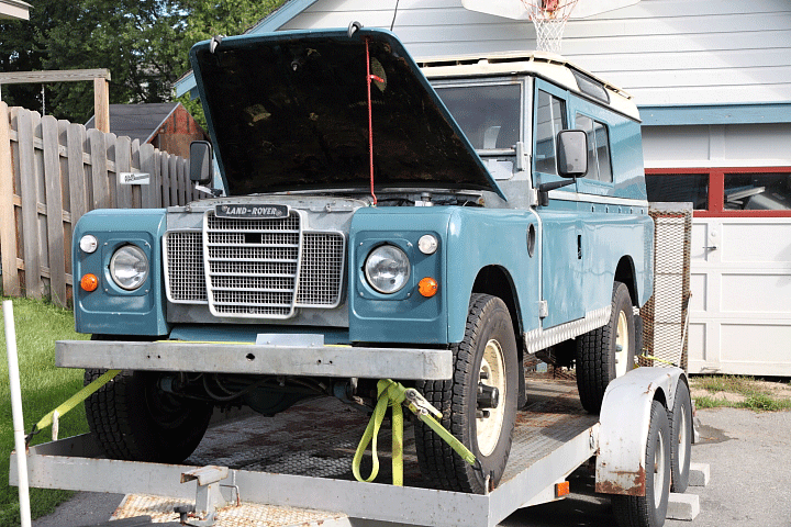 A blue-green 1979 Land rover on a trailer in a driveway with its hood up.