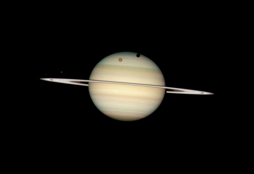 The planet Saturn, viewed nearly edge-on, with four of its moons visible in the foreground