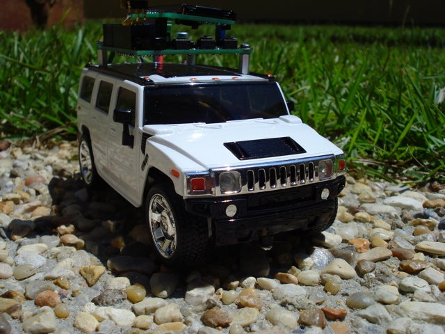 A white remote-controlled truck with an Arduino on top of it, sitting on a gravel path near a lawn.