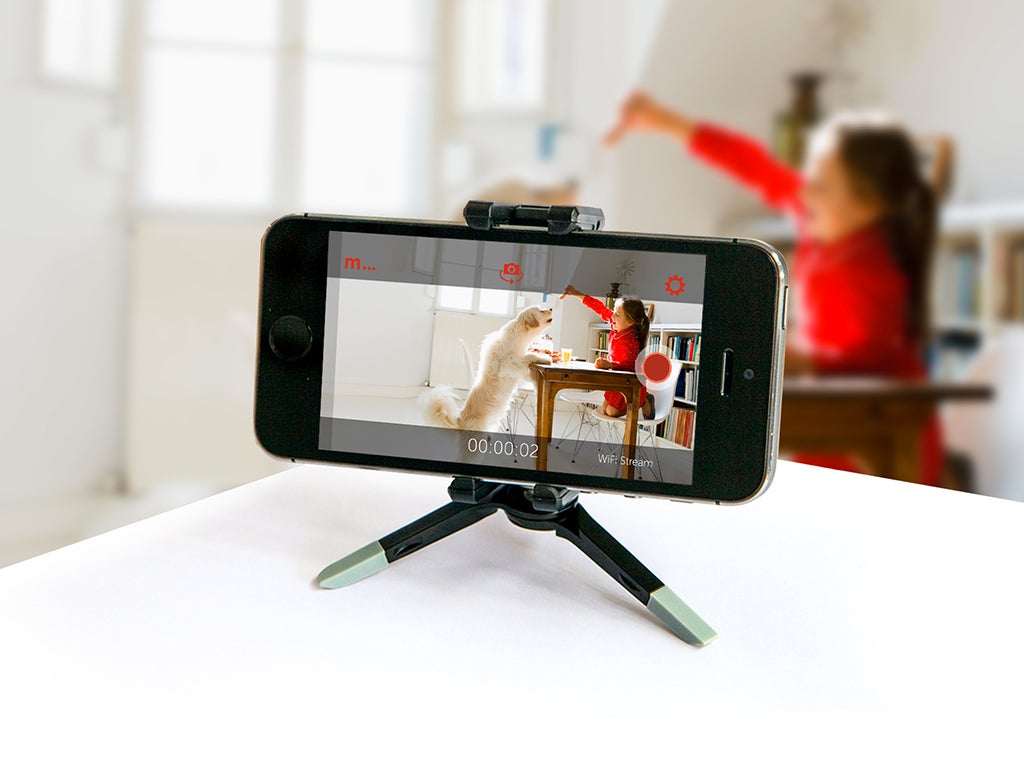 A phone set up as a home security camera on a tripod, watching a child in a red shirt playing with a white dog.