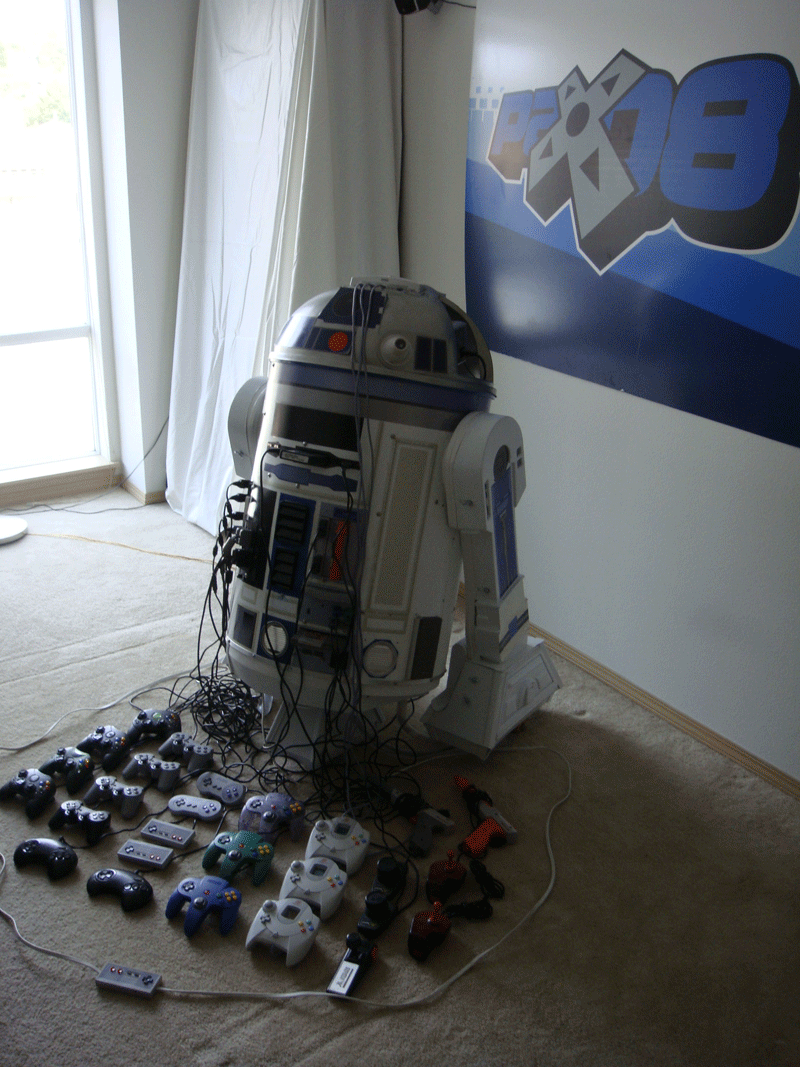 An R2-D2 cooler in the corner of a carpeted room, with video game controllers organized in front of it.