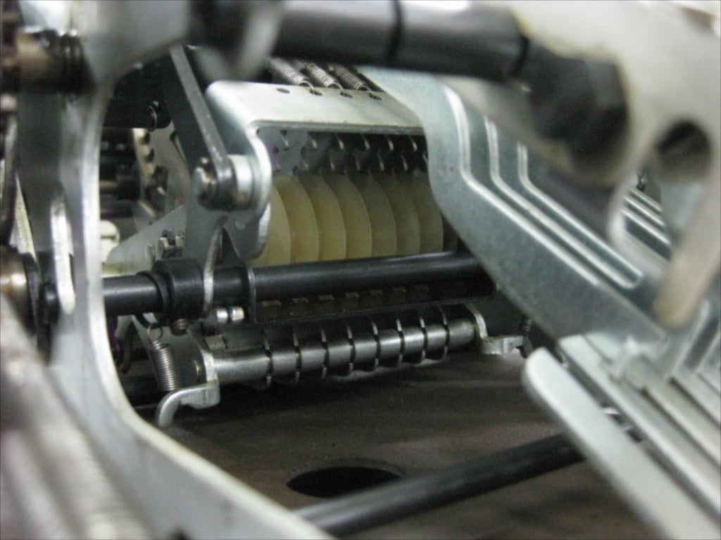 Pictured here are the counter wheels (beige plastic) and the "gears" of the previous picture (steel, barely visible, engaged with the top of the counter wheels). As those gear arms turn, they rotate the counter wheels. In using this machine, a user enters on the keypad a number to be added to the running total and pulls the lever. When the lever is pulled, the number bars are extended. The amount of their movement is limited by each digit of the number entered on the keypad. That movement is translated through a linkage into rotation of the "gear" arms pictured here, which is then translated into a rotation of the counter wheels. Because the counter wheels had an initial state (either 0, if the machine has just been zero-ed, or the previous running total), the additional rotations add the newly entered number to the running tally. The wheels have 10 positions, representing a value from 0 to 9 for each significant digit. For example, if the ones position of the current total was 2, meaning that the counter wheel is turned to the 2nd tooth already, and the number 3 is added to the total, the gear arm will be made to move 3 teeth, which will turn the ones counter wheel 3 more positions to a new total of 5.
