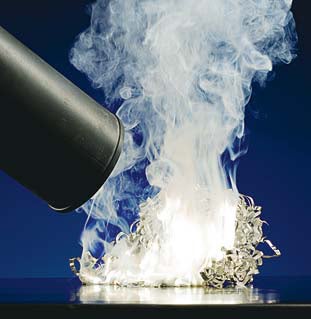 A burning pile of magnesium expelling white smoke against a dark blue background, with a fire extinguisher nozzle nearby setting it on fire.