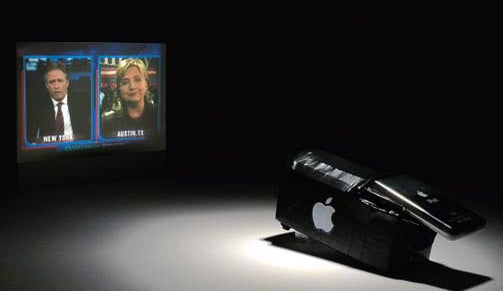 A DIY video projector made out of an iPod and a slide-projector lens projecting The Daily Show onto a black background.