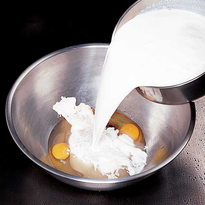 Pouring milk from a small metal container into a metal bowl with eggs in it, against a black background.