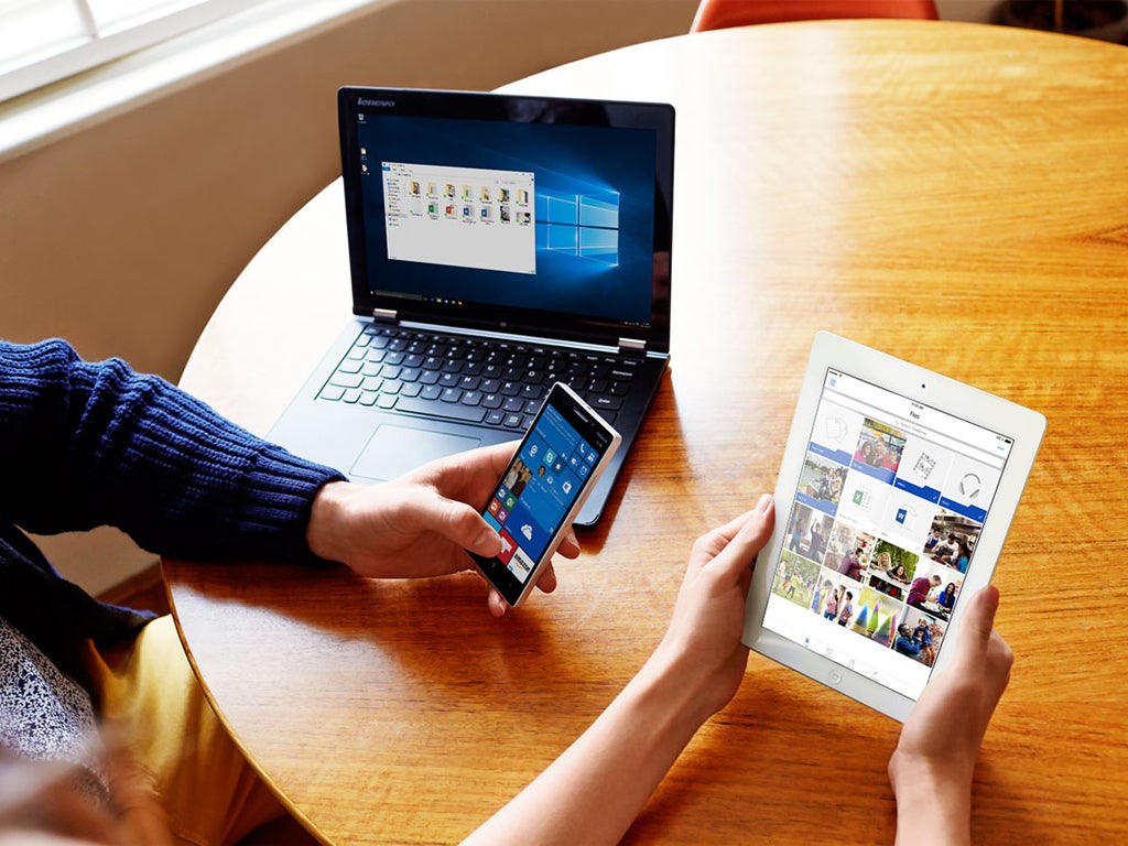 Two people using Microsoft OneDrive on a laptop, a phone, and a tablet, while sitting at a wooden table.
