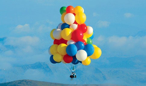 A person, John Ninomiya, suspended under a cluster of multi-colored balloons floating in front of a mountain range.