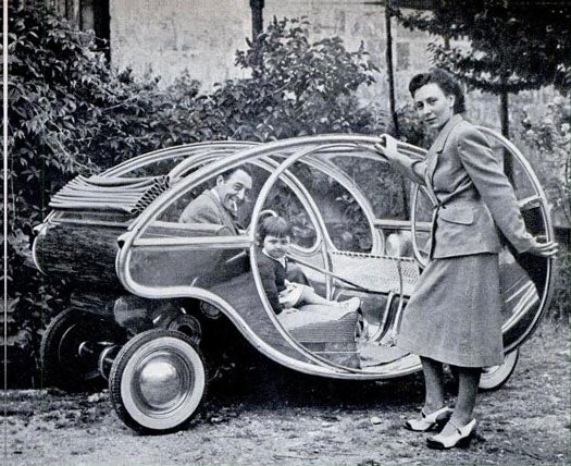 A man in an egg-shaped car with a small child in the passenger seat and a woman standing by the open passenger door.