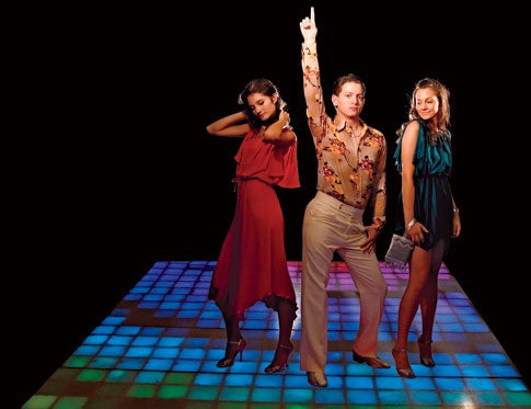 A woman in a red dress, a man in disco attire, and a woman in a green dress posing on a multi-colored disco floor.