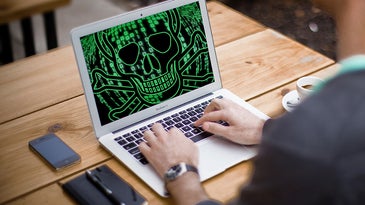 A person sitting in front of a laptop that has a skull and crossbones in green code on the screen.