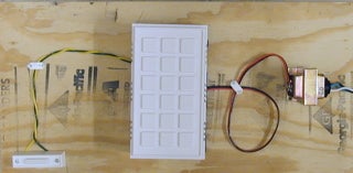 A white doorbell with wires coming out of it, on plywood.