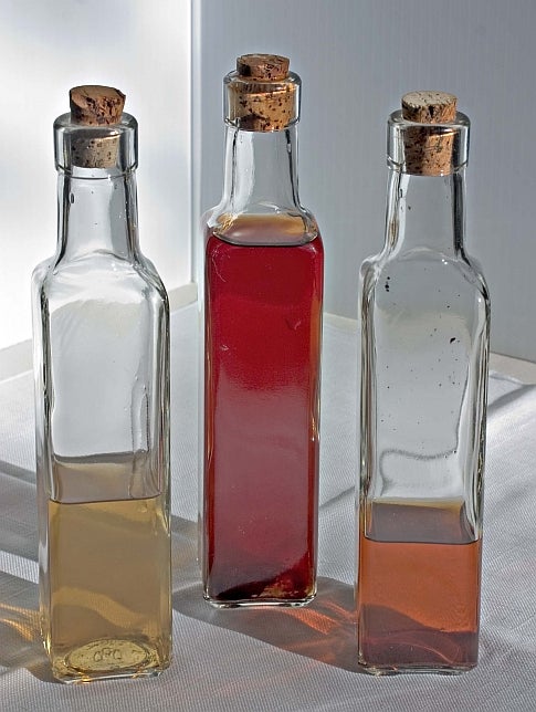 Three thin, corked rectangular bottles on a white surface. The right one is half full of yellow vinegar, the middle one nearly full of red vinegar, and the right one about a quarter full of orange vinegar.