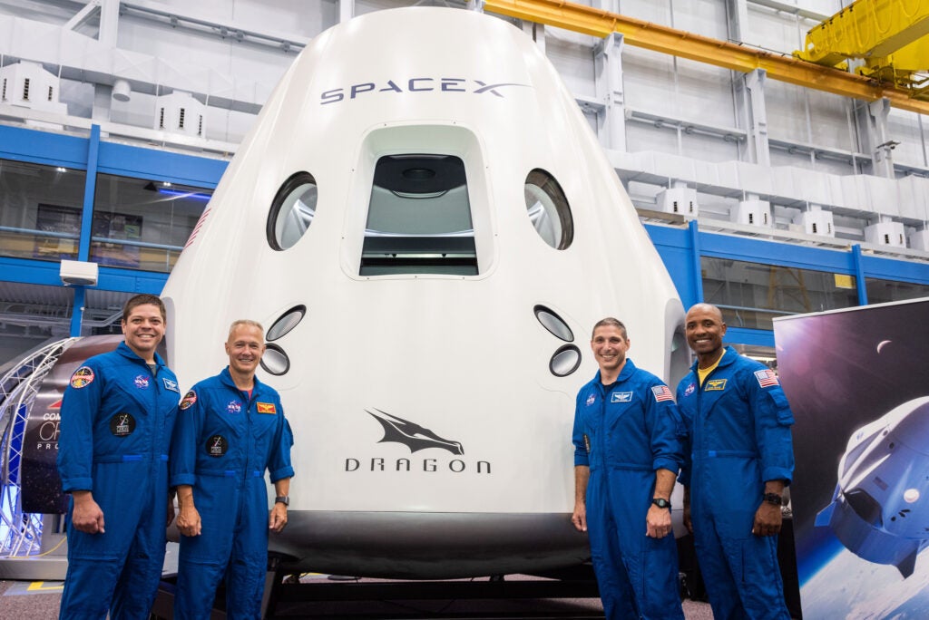 four astronauts in blue jumpsuits standing in front of dragon capsule