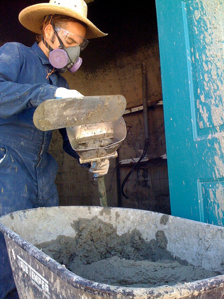 A person wearing blue coveralls and a cowboy hat reloading a tirolessa sprayer from a wheelbarrow full of concrete.