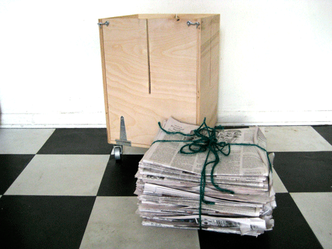 A DIY newspaper baler on a black-and-white tile floor with a stack of newspapers in front of it that's tied with green twine.