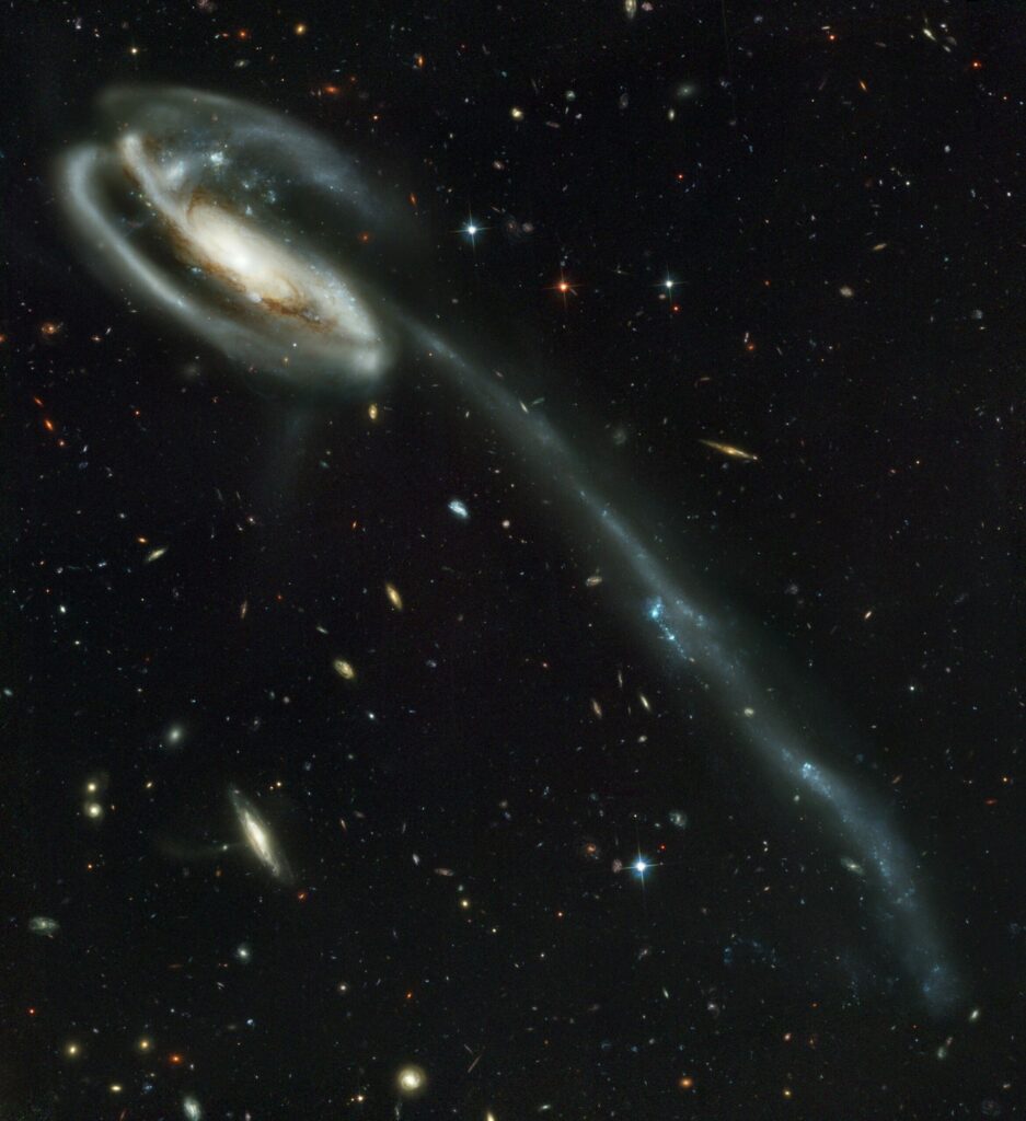 A galaxy in the upper left of the image has a huge tail that stretches down to the lower right of the image