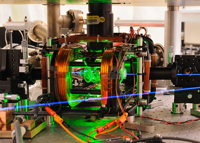 This is a composite photo of a microwave apparatus used in NIST quantum computing experiments. A pair of ions are trapped by electric fields and manipulated with microwaves inside a glass chamber at the center of the apparatus. The chamber is illuminated by a green light-emitting diode for visual effect. An ultraviolet laser beam used to cool the ions and detect their quantum state is colorized to appear blue.