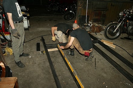 Two men working on building the frame of a pedal-powered tank, while another man stands off to the side.