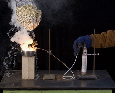 Smoke and fire from a metal bowl under a net full of popcorn in a lab setup.