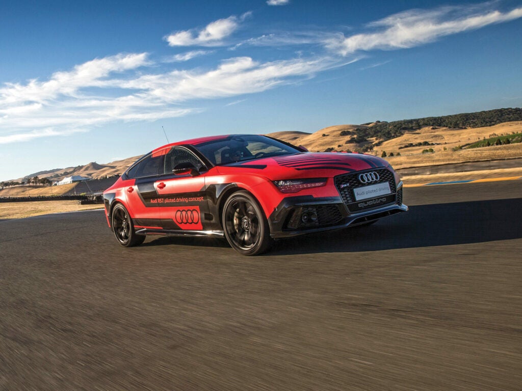 A red Audi sports car with black racing stripes, driving on a desert road under a clear blue sky